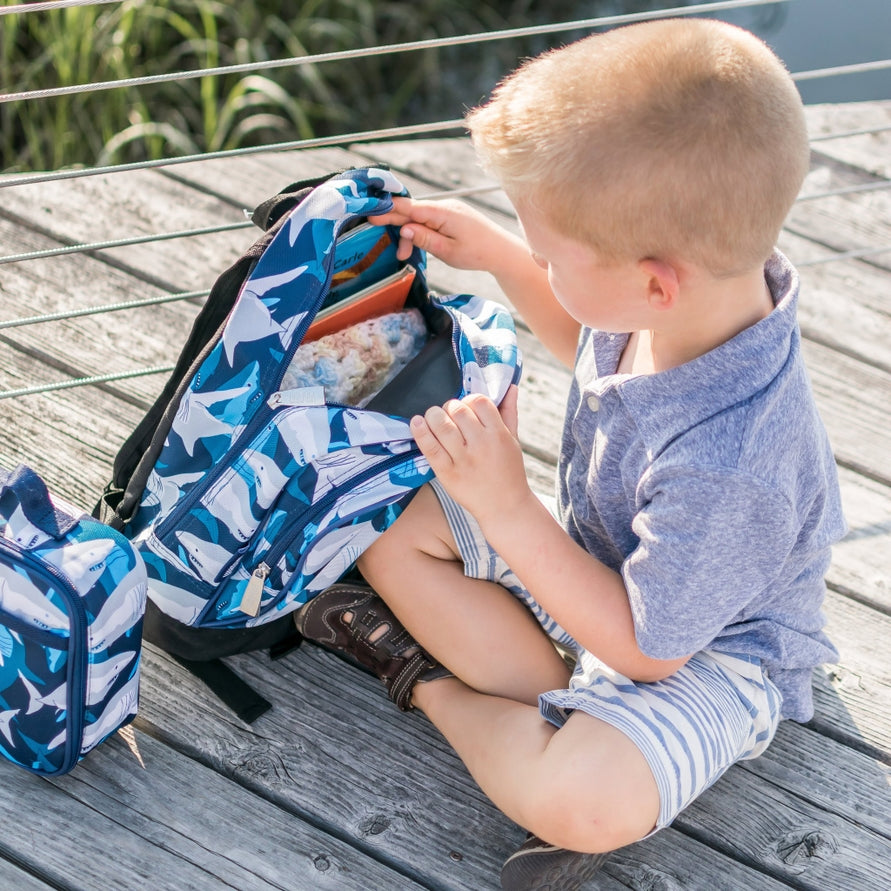 A young boy sits on a wooden dock, opening a blue and white shark-patterned backpack next to a matching lunch bag.