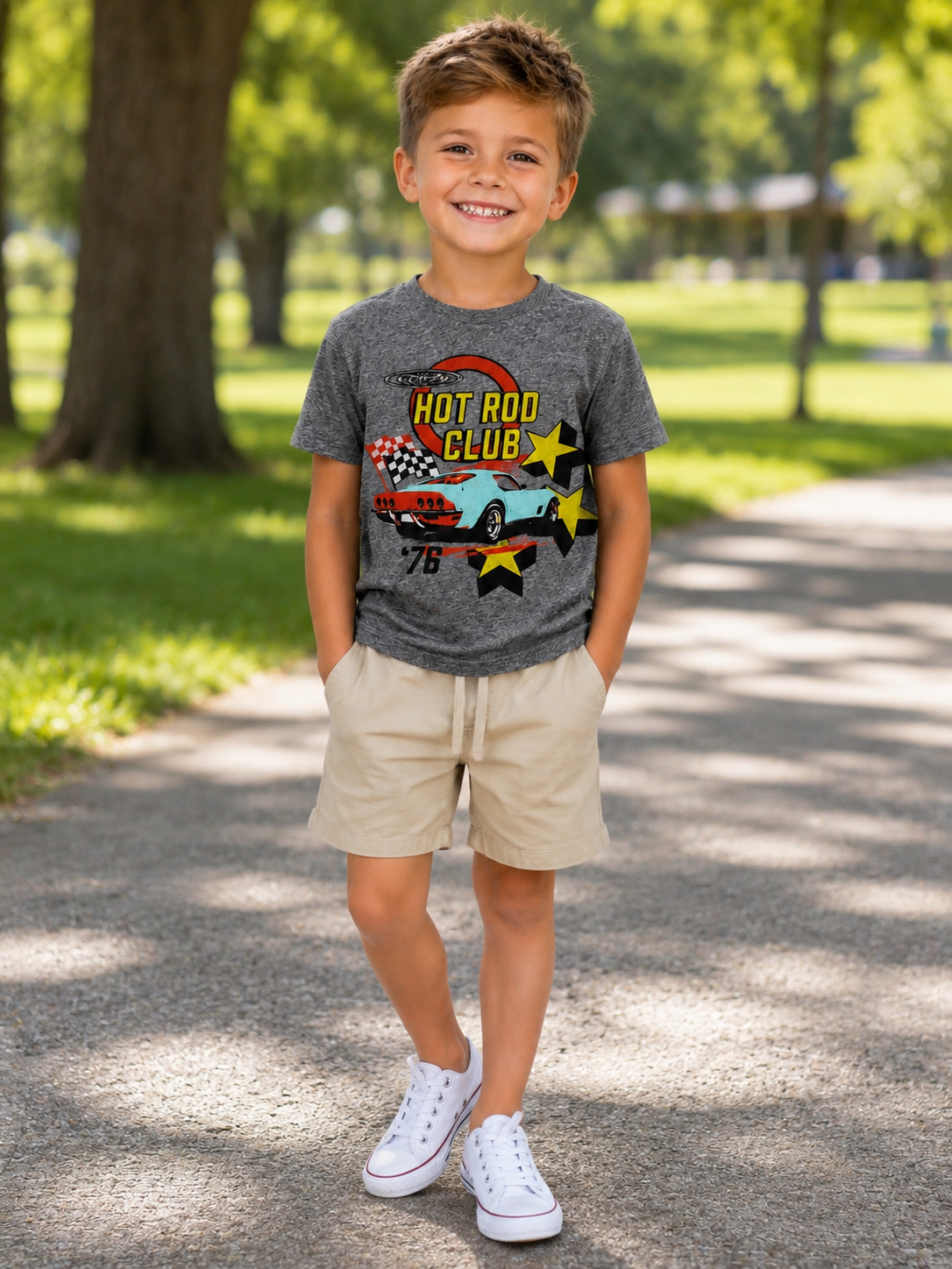 A young boy stands on a paved path in a sunny park, wearing a heather gray graphic t-shirt with a 'Hot Rod Club' race car design, khaki shorts, and white sneakers.