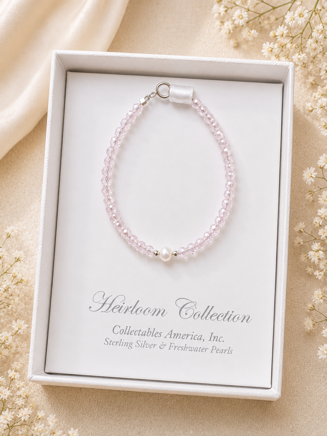 Pink beaded bracelet in a white box with 'Heirloom Collection' text, surrounded by small white flowers.