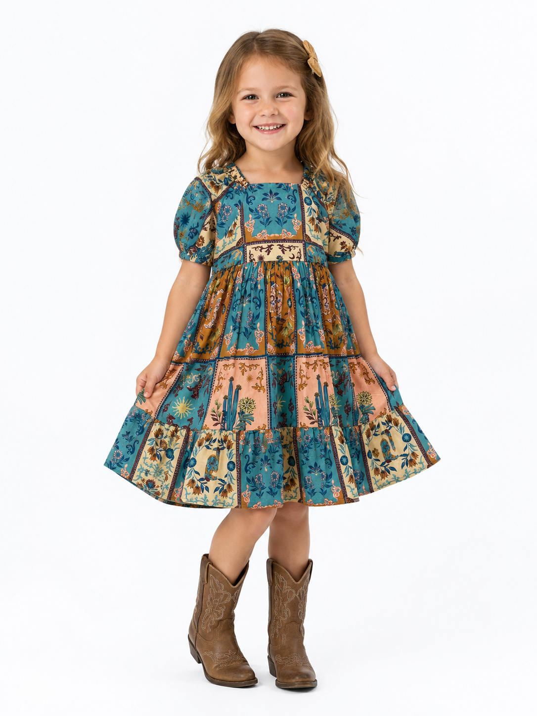 A young girl with long light brown hair stands against a plain white background wearing a tiered western-style dress with a blue, tan, and brown patchwork print, short puff sleeves, and brown cowboy boots.
