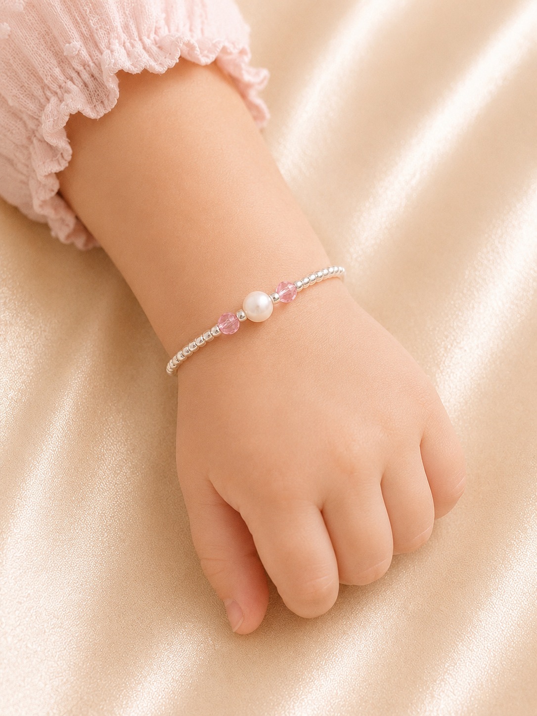 Child's hand wearing a bracelet with a pearl and pink gemstone on a soft beige background