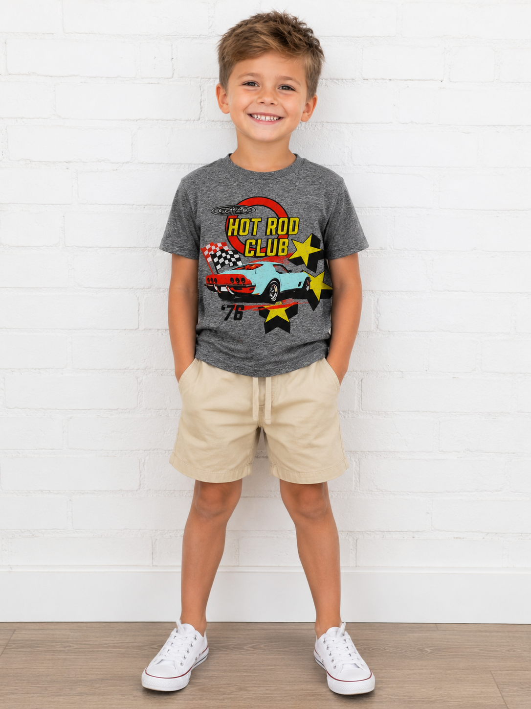 A young boy stands against a white brick wall wearing a heather gray graphic t-shirt with a hot rod car design, beige shorts, and white sneakers.