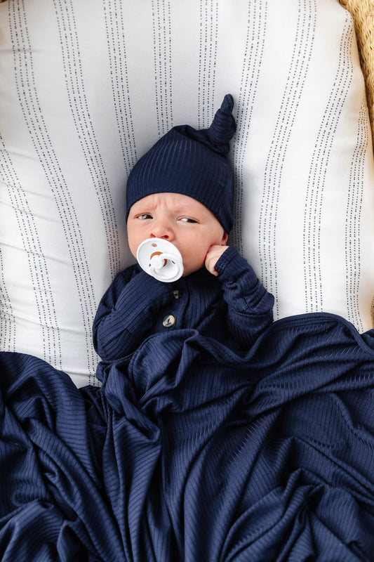 Newborn baby wrapped in a navy blue blanket and wearing a matching navy blue hat with a white pacifier.