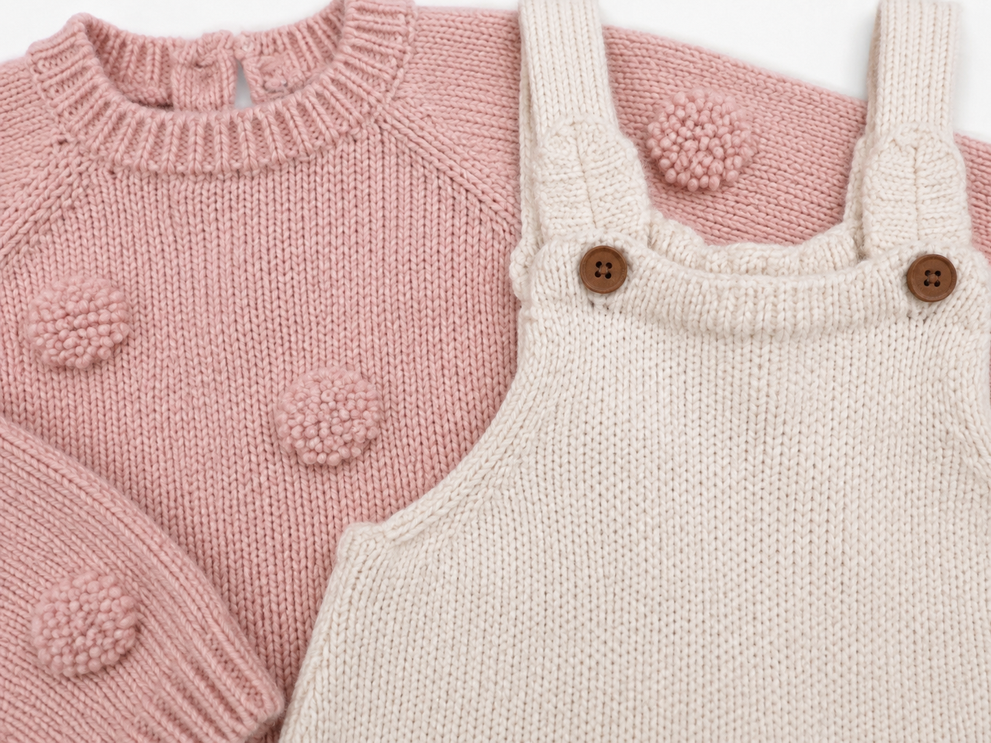 A close-up of a knitted baby outfit featuring a dusty pink sweater with pom-pom details and an off-white knit overall with brown buttons, layered together on a white background.