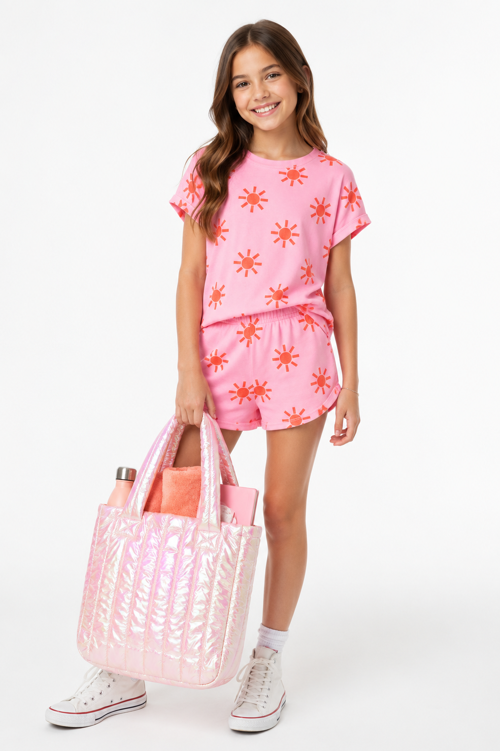 A young girl stands against a white studio background wearing a pink short-sleeve t-shirt and matching shorts with orange starburst patterns, holding a large iridescent pink tote bag.