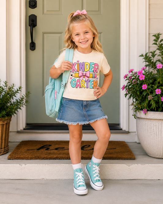 A young girl stands on a front porch wearing a cream graphic T-shirt that says 'KINDER GARTEN' in colorful letters, a denim skirt, mint socks, mint sneakers, and a mint backpack.
