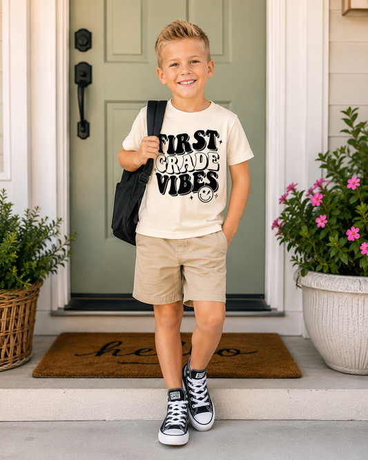 A young boy stands on a front porch wearing a cream graphic t-shirt that reads 'FIRST GRADE VIBES' in black, khaki shorts, and black-and-white sneakers, with a black backpack strap over his shoulder.