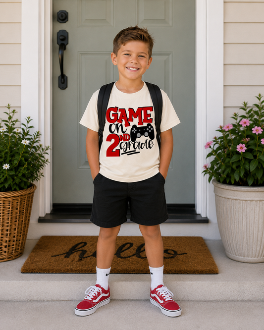 A young boy stands on a front porch wearing a cream graphic T-shirt that reads 'GAME OR 2nd gradle' in red and black, black shorts, white socks, and red sneakers. He has a black backpack on.
