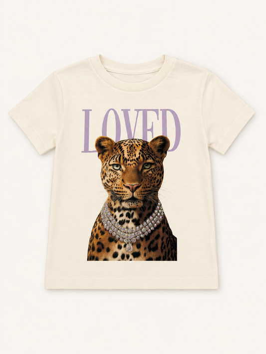 An off-white short-sleeve t-shirt featuring a graphic print of a leopard wearing a diamond necklace, with the word 'LOVED' printed in a lavender-purple serif font above it.