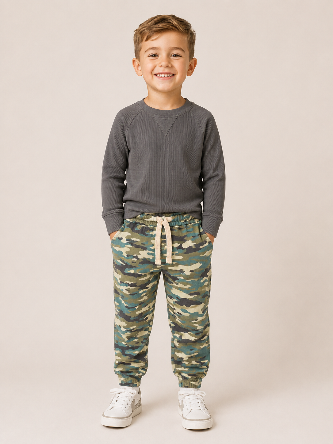 A young boy smiling and standing against a plain beige studio background, wearing a dark gray long-sleeve sweatshirt, camouflage print jogger pants with a cream drawstring, and white sneakers.